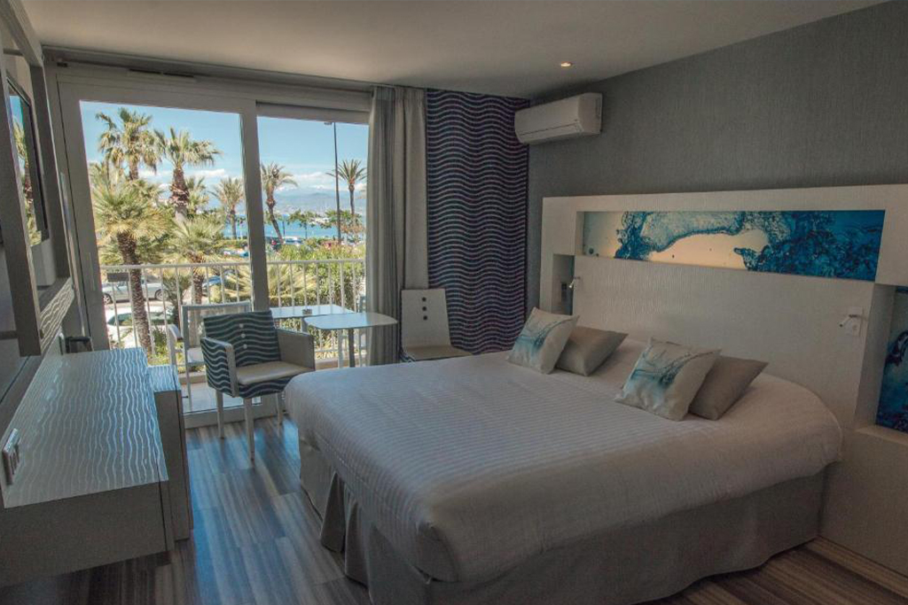 Hotel Josse room with a large bed, desk area, and balcony overlooking palm trees and the Mediterranean.