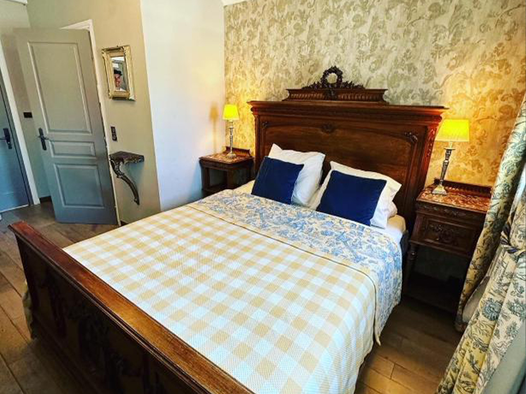 Hotel Le Ponteil room with a wooden bed, checkered bedspread, bedside lamps, and warm decorative wallpaper.
