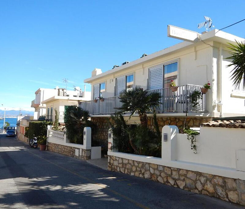 Exterior view of Hotel Miramar in Antibes, a white Mediterranean building facing the sea with palm trees and a stone wall.