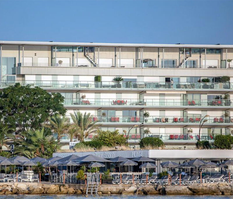 Exterior of Hotel Royal Antibes, a modern seafront building with balconies, glass railings, and surrounding palm trees.